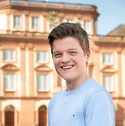 Georg Schade stands in front of Mannheim Palace, laughing. He has short brown hair and is wearing a light blue long-sleeved shirt.