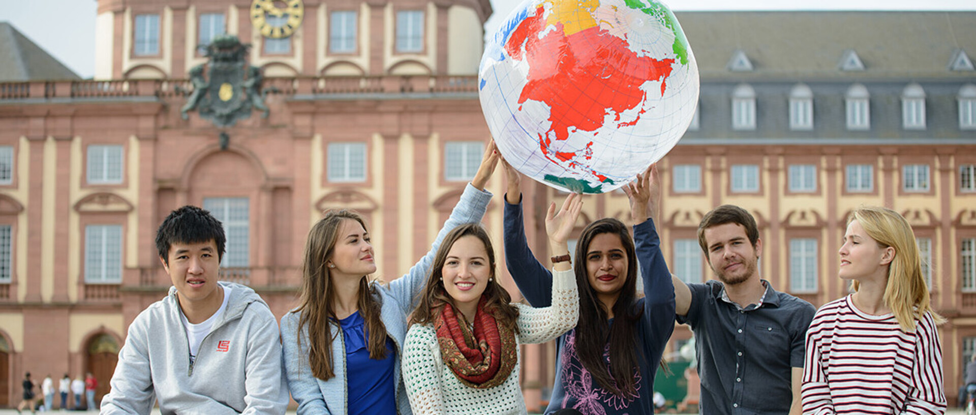 Sechs Studierende sitzen auf einer Bank im Ehrenhof und halten eine Weltkugel hoch.