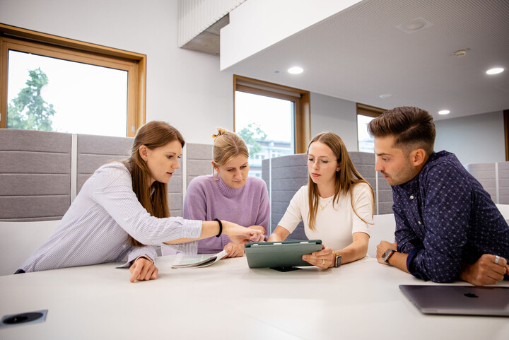 Vier Studierende sitzen in einem hellen Seminarraum über ein Tablet gebeugt.