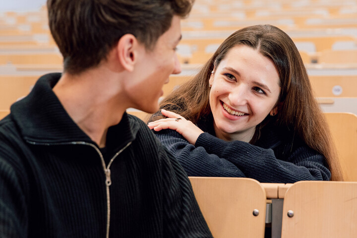 Eine Studentin und ein Student sitzen in einem Hörsaal und unterhalten sich. Sie lächelt.