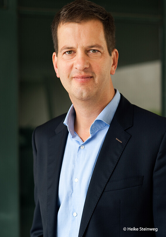 Philipp Lepenies trägt ein hellblaues Hemd und eine schwarze Anzugjacke. Das Hemd ist am Kragen geöffnet, die Jacke ist offen. Er hat kurzes, braunes Haar und zeigt ein angedeutetes Lächeln. 