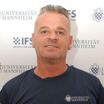 Naim Krraba hat kurze graue Haare. Er trägt ein dunkelblaues T-Shirt mit der Aufschrift "Universität Mannheim"
