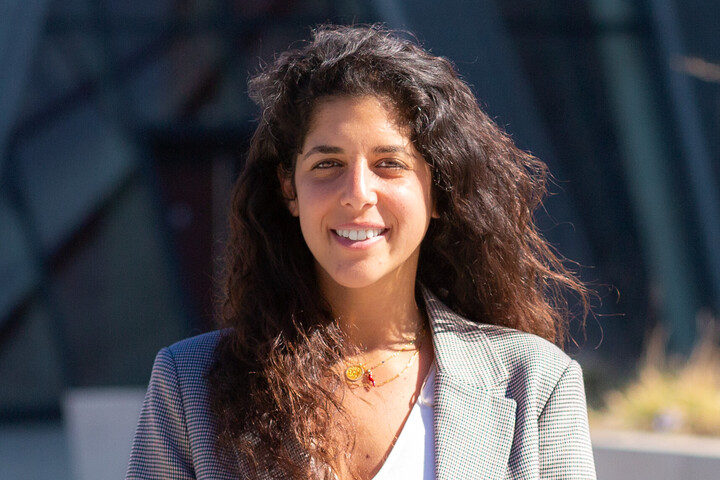 Alumna Professorin Hana Attia hat lange braune, gelockte Haare, trägt einen grauen Blazer und lächelt freundlich.