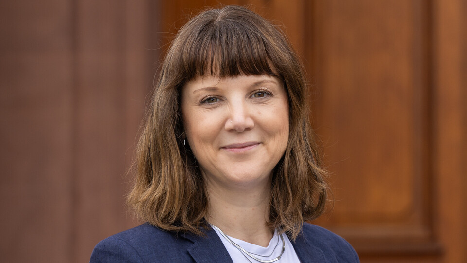 Frau mit schulterlangem braunem Haar trägt weißes T-Shirt und dunkelblauen Blazer vor Holzwand