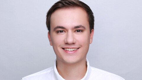 Bastian Koch. He is wearing a white shirt and is standing in front of a grey photowall.