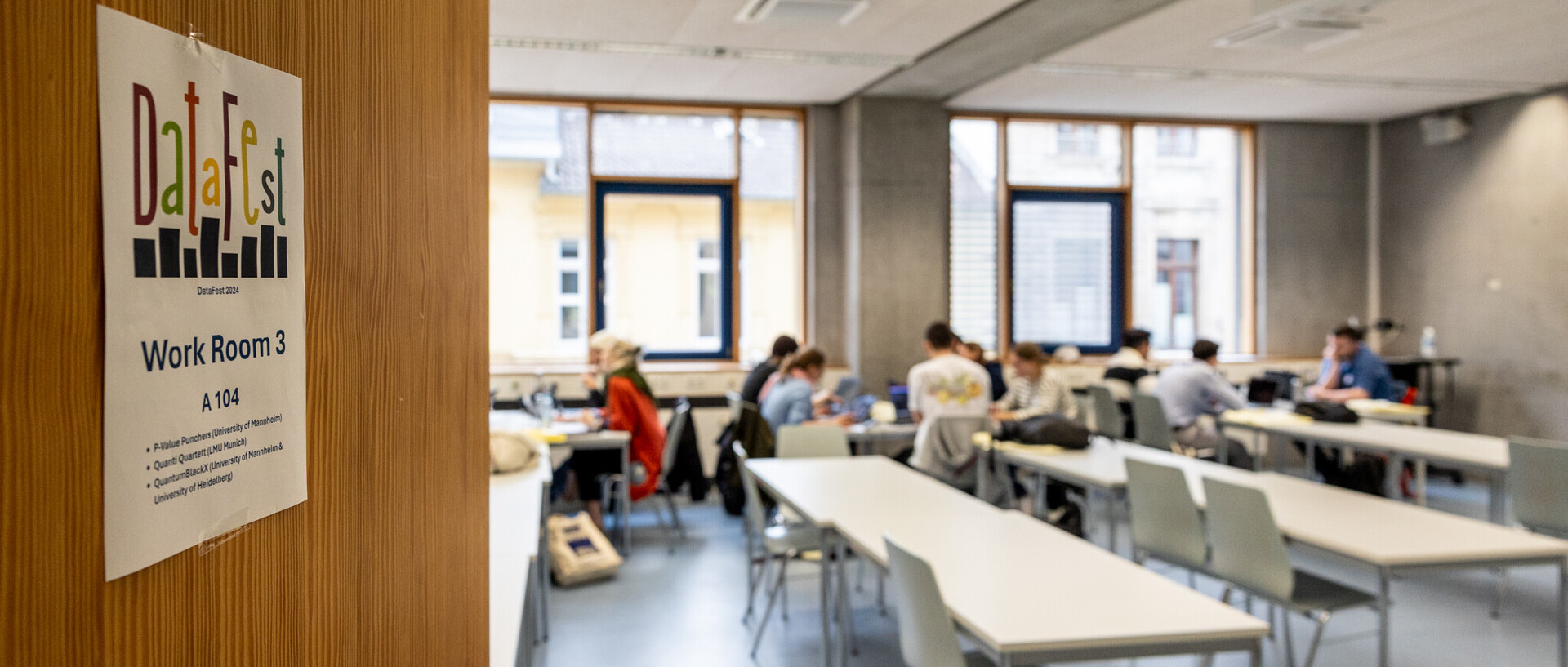 Raum mit arbeitendenden Studierenden mit Blick durch die Tür des Raums auf der ein Plakat mit dem Logo des DataFests befestigt ist.. 