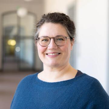 Portraitaufnahme Simone Fényes, sie hat dunkle zusammengebundene Haare, trägt eine Brille, einen blauen Pullover und lächelt freundlich in die Kamera