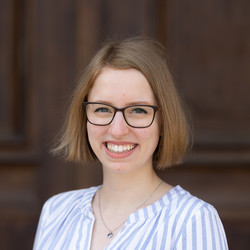 Paula Riese has short blonde hair and laughs into the camera. She wears glasses and a blue and white striped blouse.