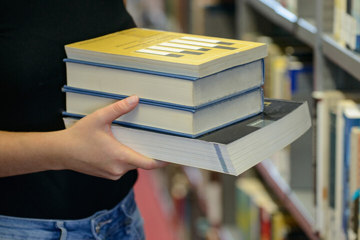 Eine Person hält Bücher in der Hand und im Hintergrund ist ein Bücherregal der Universitätsbibliothek zu sehen.