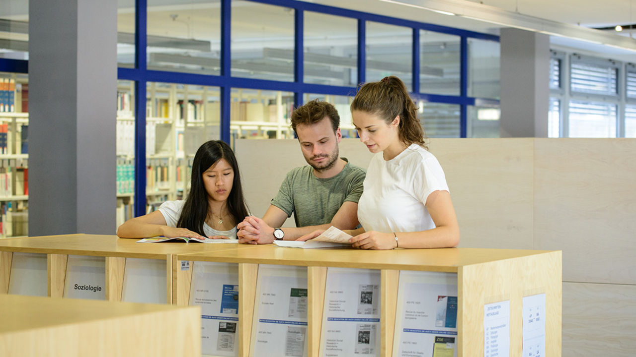 Zwei Studentinnen und ein Student stehen in der Bibliothek an einem Regal. Sie tragen T-Shirts und halten weiße Blätter in den Händen.