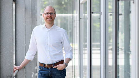 Eine Person trägt ein weißes Hemd sowie eine blaue Jeans und steht in einem Flur. Die Person heißt Jochen Gebauer.