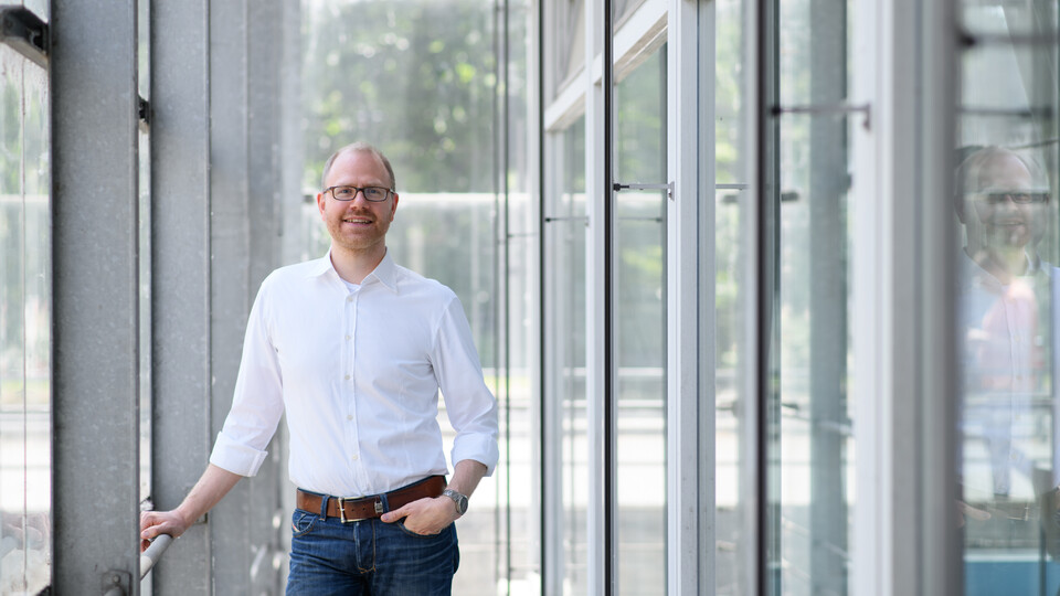 Eine Person trägt ein weißes Hemd sowie eine blaue Jeans und steht in einem Flur. Die Person heißt Jochen Gebauer.