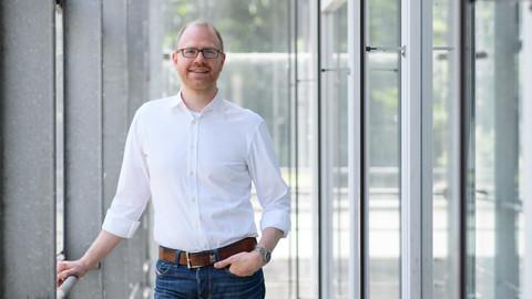 Eine Person trägt ein weißes Hemd sowie eine blaue Jeans und steht in einem Flur. Die Person heißt Jochen Gebauer.