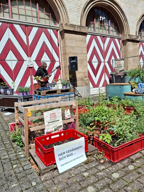 Mann mit Gitarre und Pflanzen davor in rotem Plastik-Behälter vor Haus mit großen weiß-rot-gestreiften Holztüren
