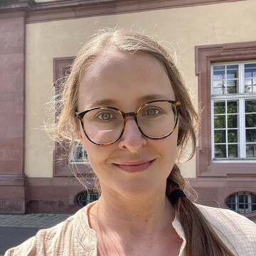 Frau mit langem, hellbraunem Haar, das zu einem Zopf gebunden ist, trägt eine beige Bluse mit Knöpfen, steht vor Mannheimer Schloss mit großen Fenstern