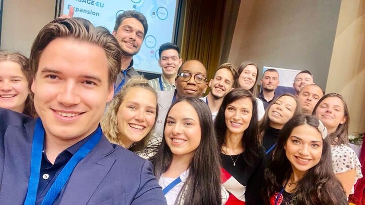 Selfie von 16 Personen in schicker Kleidung. Im Hintergrund ist ein Bildschirm mit den Worten "Engage.EU".