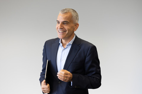 Thomas Fetzer trägt ein weißes Hemd sowie einen schwarzen Blazer und steht vor einer weißen Wand. Er hält ein Tablet unter seinem Arm.