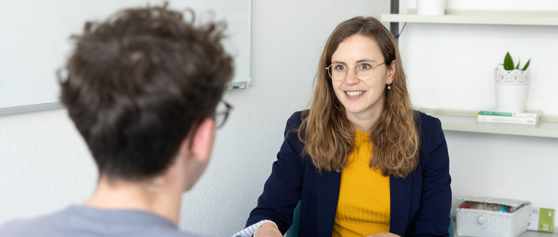 Leonie Schültke hat lange hellbraune Haare. Sie trägt eine Brille mit einem dünnen Rahmen und ein gelbes Oberteil mit einem dunkelblauen Blazer. Schültke sitzt an einem Tisch in einem Büro und gegenüber von ihr sitzt eine weitere Person.