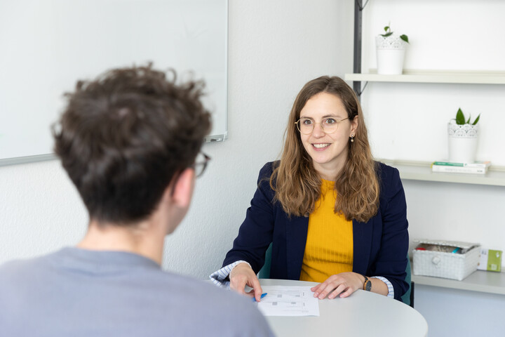 Leonie Schültke hat lange hellbraune Haare. Sie trägt eine Brille mit einem dünnen Rahmen und ein gelbes Oberteil mit einem dunkelblauen Blazer. Schültke sitzt an einem Tisch in einem Büro und gegenüber von ihr sitzt eine weitere Person.