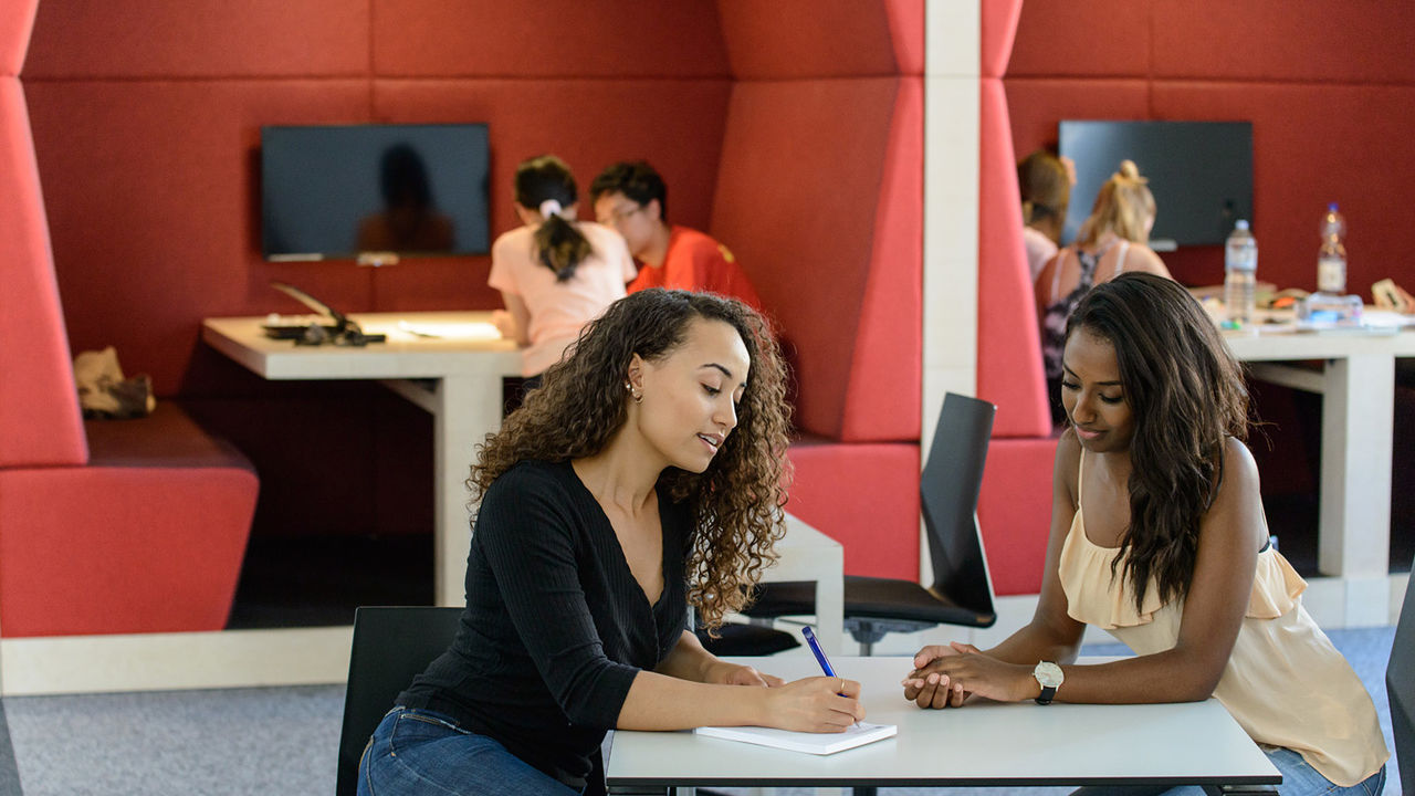 Eine Studentin schreibt etwas auf ein Papier. Eine weitere Studentin schaut interessiert dabei zu.