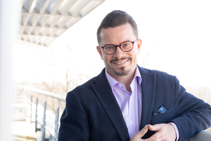 Portrait von Nicolas R. Ziebarth. Er trägt Brille, ein fliederfarbenes Hemd und ein dunkelblaues Sakko.
