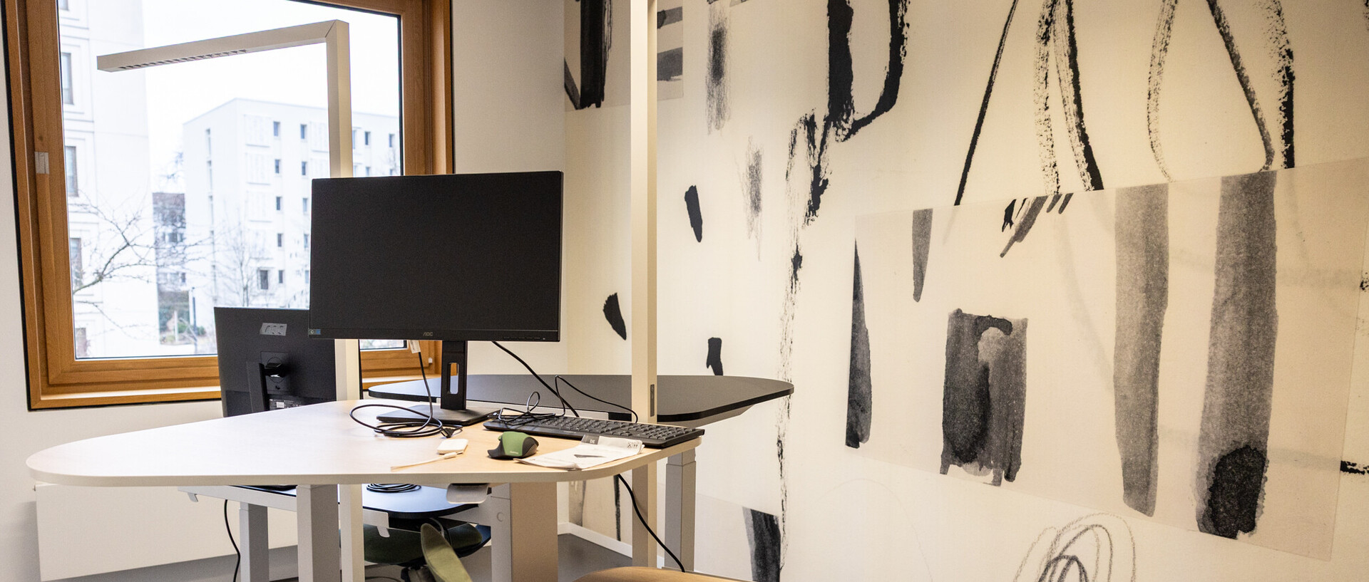 Auf dem Foto blickt man in einen CAS Raum Richtung Fenster. Davor stehen zwei höhenverstellbare Tische mit Bildschirmen darauf. An der Wand rechts ist eine Tapete mit schwarz-weiss Grafik angebracht.