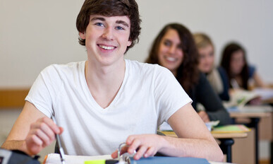 Student working on homework in classroom Ein Student sitzt an einem Tisch und lächelt. Er hat braunes Haar und trägt ein weißes T-Shirt. Vor ihm liegt ein Ordner.