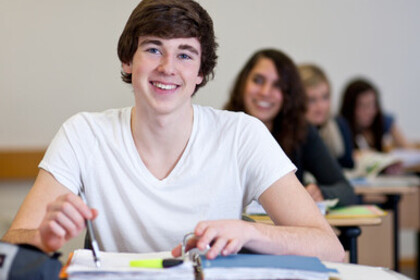 Ein Student sitzt an einem Tisch und lächelt. Er hat braunes Haar und trägt ein weißes T-Shirt. Vor ihm liegt ein Ordner.