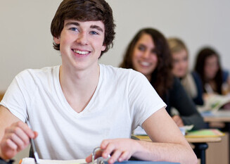 Ein Student sitzt an einem Tisch und lächelt. Er hat braunes Haar und trägt ein weißes T-Shirt. Vor ihm liegt ein Ordner.