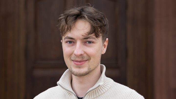 Tobias Großbölting. He is wearing a white sweater and is standing in front of a brown door.