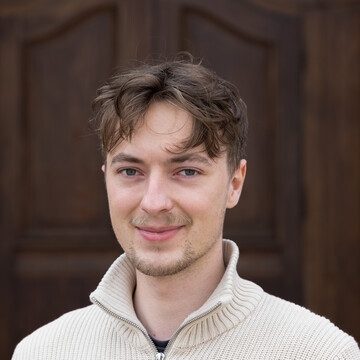 Tobias Großbölting. Er trägt einen weißen Pullover mit hohen Kragen und steht vor einer Holztür.