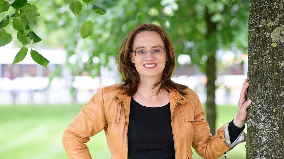 Portrait von Michele Tertilt. Sie trägt ein schwarzes Oberteil sowie eine orange farbige Jacke. Sie steht auf einer Grünfläche und hebt ihre Hand an einen Baum.
