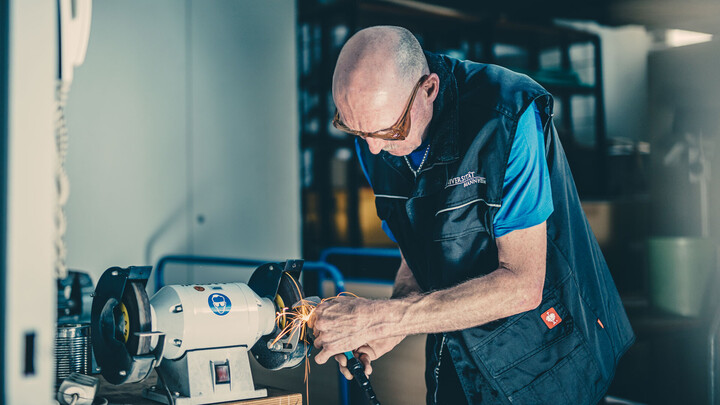 Mitarbeiter mit Schutzbrille schleift Metall an einer Werkbank, Funken fliegen.