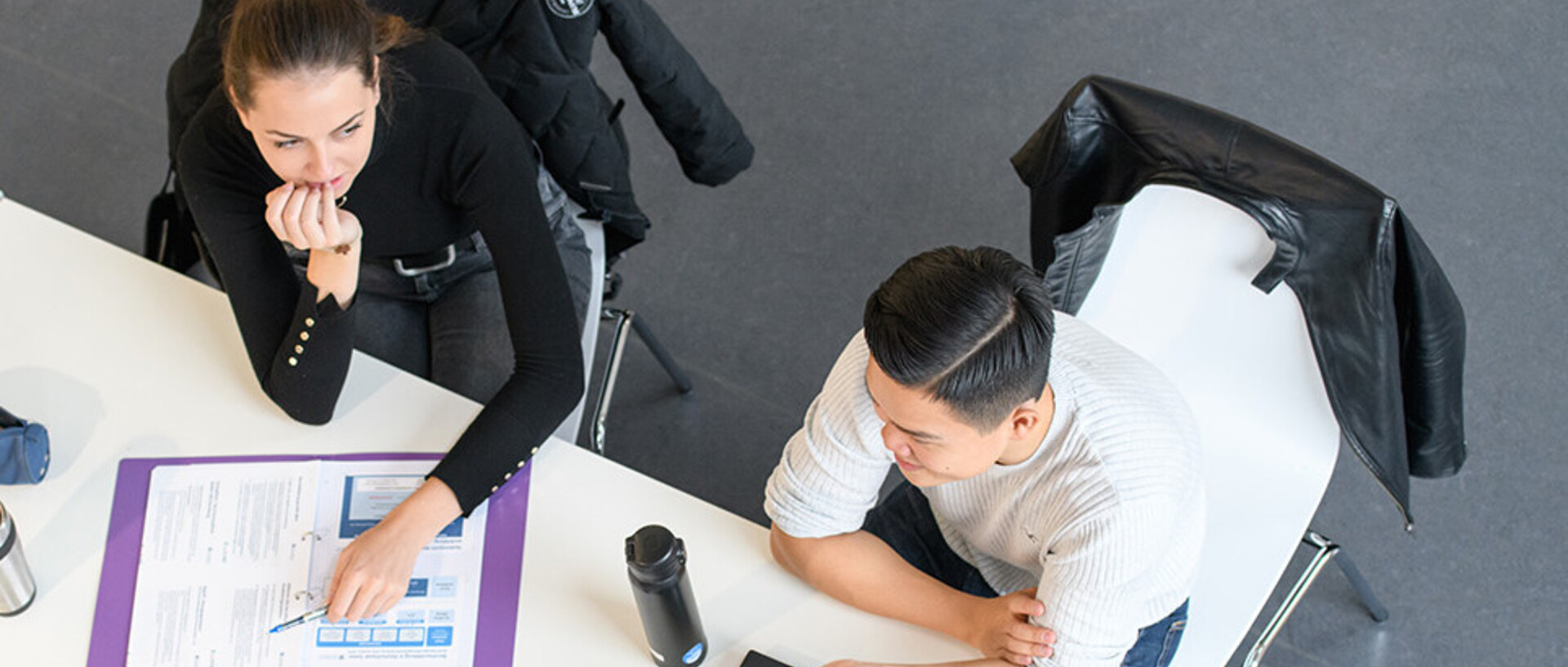 Zwei Studentinnen und ein Student sitzen an einem Tisch und unterhalten sich. Die Vogelperspektive zeigt den Tisch von oben, auf dem ein Ordner, Getränke und ein Laptop liegen.