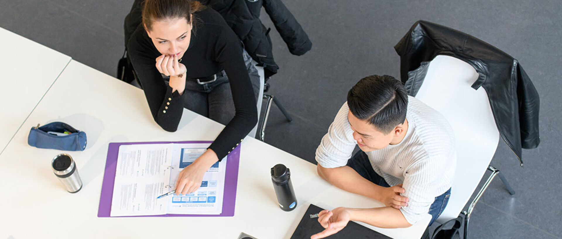 Zwei Studentinnen und ein Student sitzen an einem Tisch und unterhalten sich. Die Vogelperspektive zeigt den Tisch von oben, auf dem ein Ordner, Getränke und ein Laptop liegen.