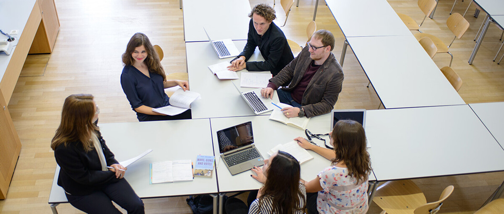 Eine Gruppe von Personen sitzt gemeinsam an Tischen und führen ein Gespräch. Aufgeklappte Laptops liegen auf den Tischen.