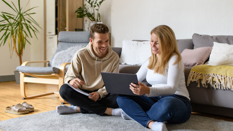 Eine Studentin und ein Student sitzen vor einem Laptop auf einem Teppich