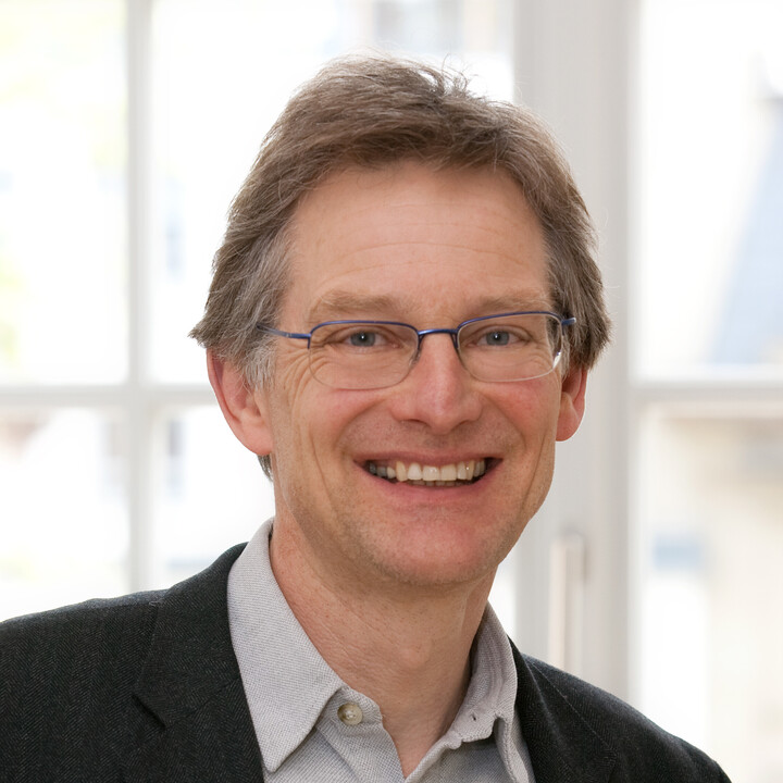 Personenportrait. Ein Mann steht vor einer Fensterfront und blickt lächelnd in die Kamera.