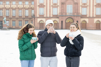 Drei Studierende stehen auf dem Ehrenhof, tragen Winterjacken und halten jeweils eine Tasse in den Händen. 