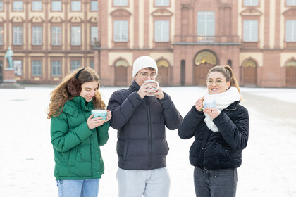 Drei Studierende stehen auf dem Ehrenhof, tragen Winterjacken und halten jeweils eine Tasse in den Händen. 