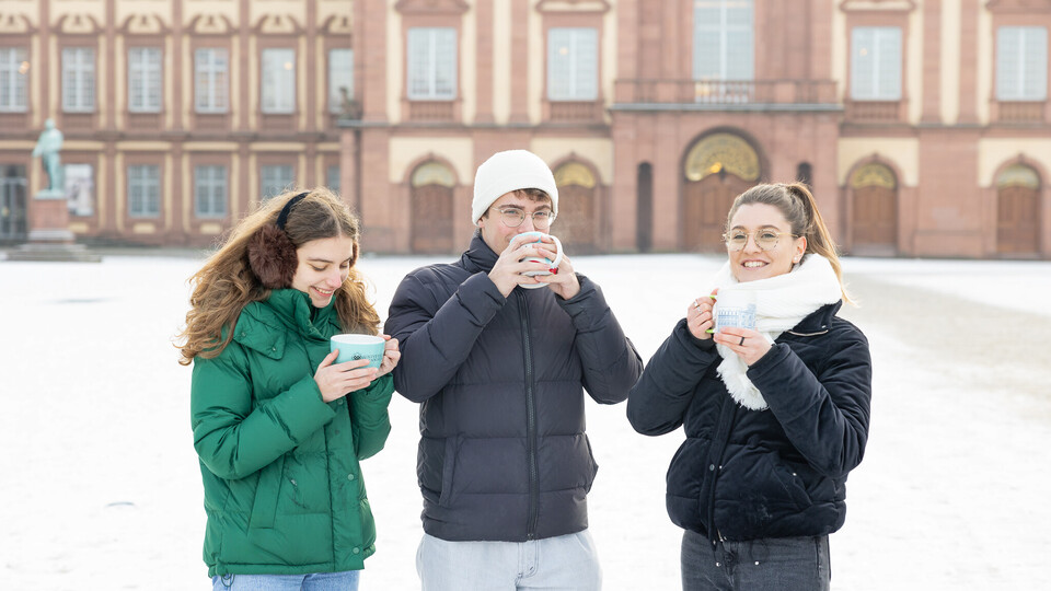 Drei Studierende stehen auf dem Ehrenhof, tragen Winterjacken und halten jeweils eine Tasse in den Händen.