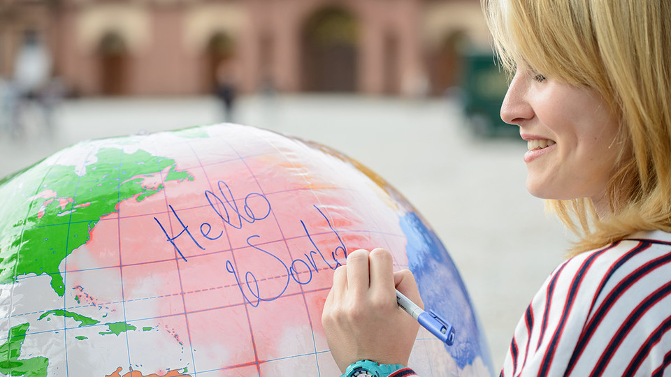 Lächelnde Frau in einem gestreiften Shirt auf dem Ehrenhof, schreibt mit einem Stift auf einen großen Globus "Hello World".
