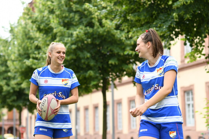 Zwei Rugby-Sportlerinnen tragen blaue Trikots und lachen gemeinsam im Grünen.