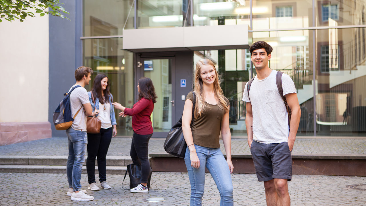 Eine Studentin und ein Student stehen vor einem gläsernen Eingang der Universität Mannheim und lächeln in die Kamera. Im Hintergrund stehen drei weitere Studierende und unterhalten sich.
