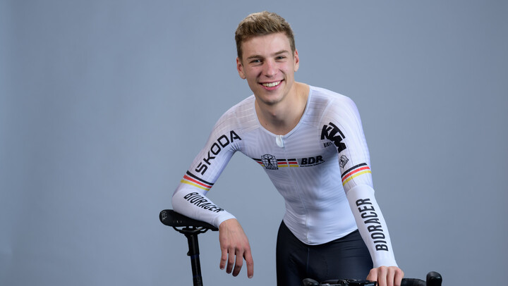 Tobias Buck-Gramcko steht vor einer grauen Wand und lächelt in die Kamera. Er trägt ein weißes Trikot und stützt sich auf seinem Rennrad ab.