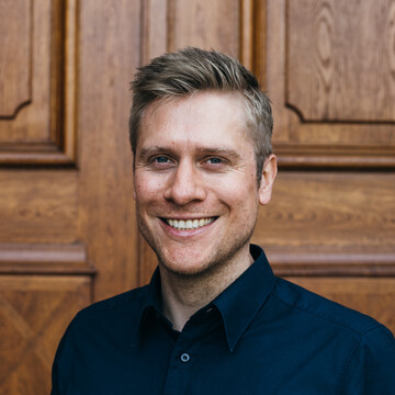 Daniel Lehnert hat kurze blonde Haare. Er trägt ein dunkles Hemd und lächelt in die Kamera.