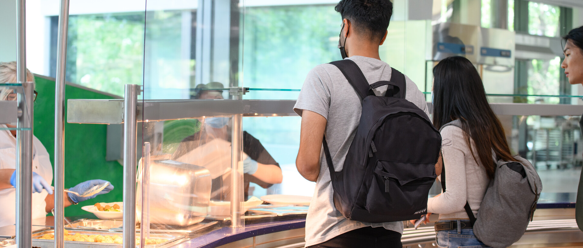 Studierende stehen vor der Essensausgabe in der Mensa und warten auf ihr Mittagessen.