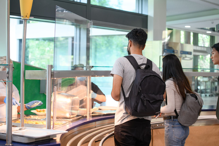 Studierende stehen vor der Essensausgabe in der Mensa und warten auf ihr Mittagessen.