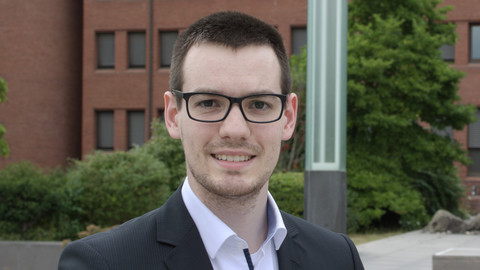 Stefan Weck. He is wearing a black suit and is standing outside in front of brown building and trees.