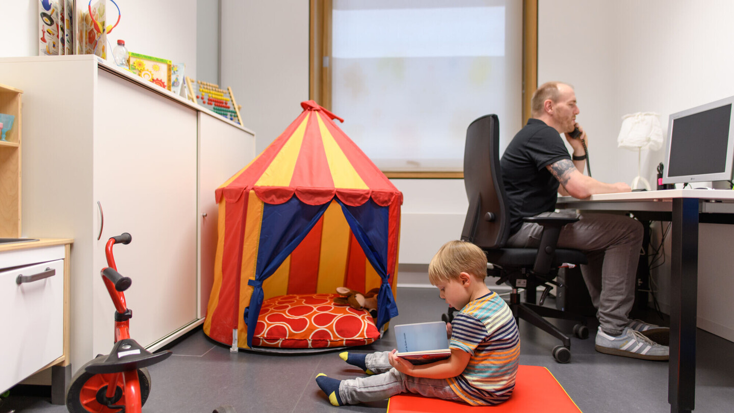 Ein Mitarbeitender telefoniert im Büro, neben ihm auf dem Boden liest sein Sohn ein Buch.